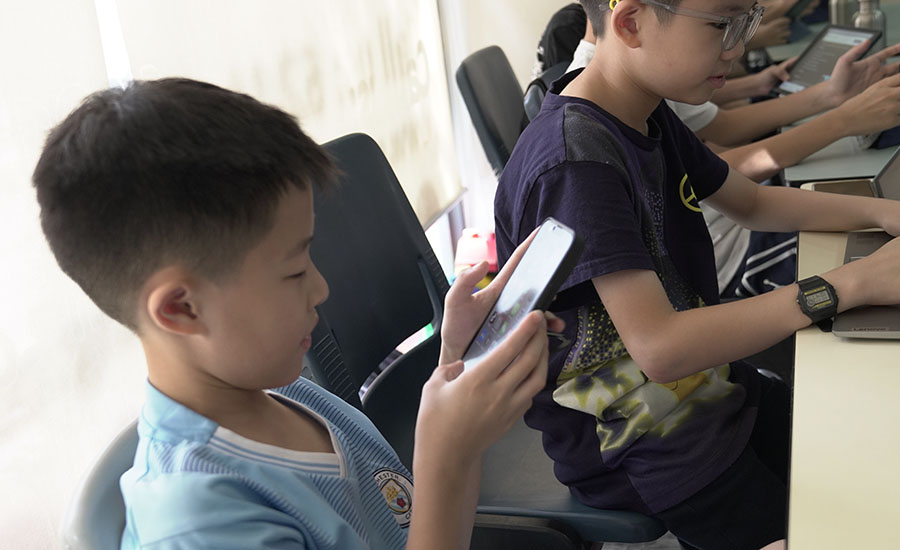 Two young boys seated at desks, one holding phone, other using laptop