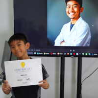 Young boy holds certificate, smiling beside screen showing smiling adult