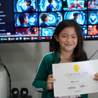 Young girl holds certificate, smiling in front of large video monitor display