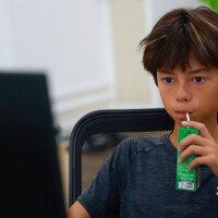Young boy sips green drink while seated at computer desk