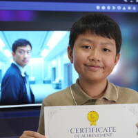 Young boy holds certificate, smiling in front of TV screen