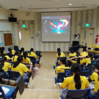 Students in yellow shirts sit in a classroom with laptops and a projector screen
