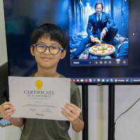 Young boy holds certificate, smiling in front of large screen displaying movie scene