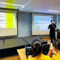 Presenter speaks to audience in classroom with two large screens displaying presentations